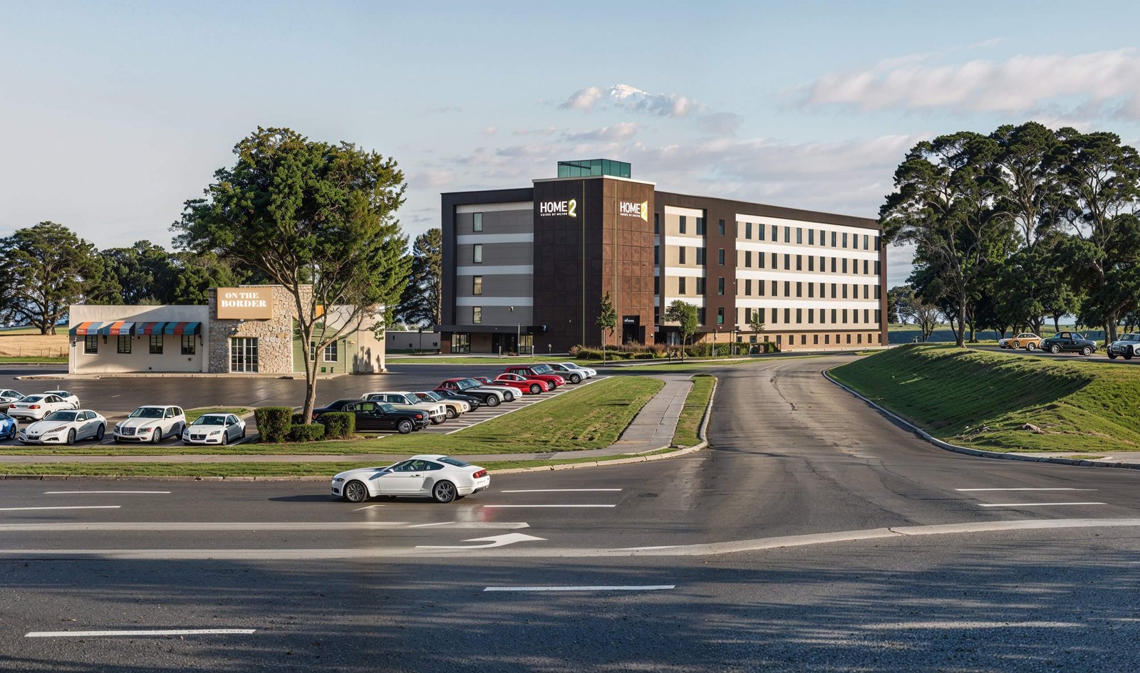 Street-level perspective of modern multi-story motel building with driveway, vehicle and landscaping in natural light