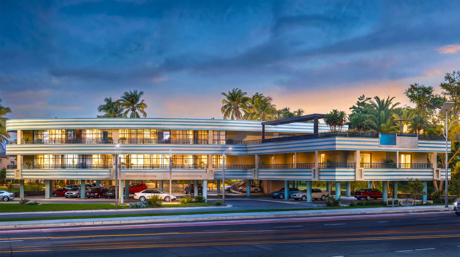 Modern apartment building with curved architecture, elevated structure, open parking, landscaped surroundings, and warm evening lighting in a contemporary residential development.